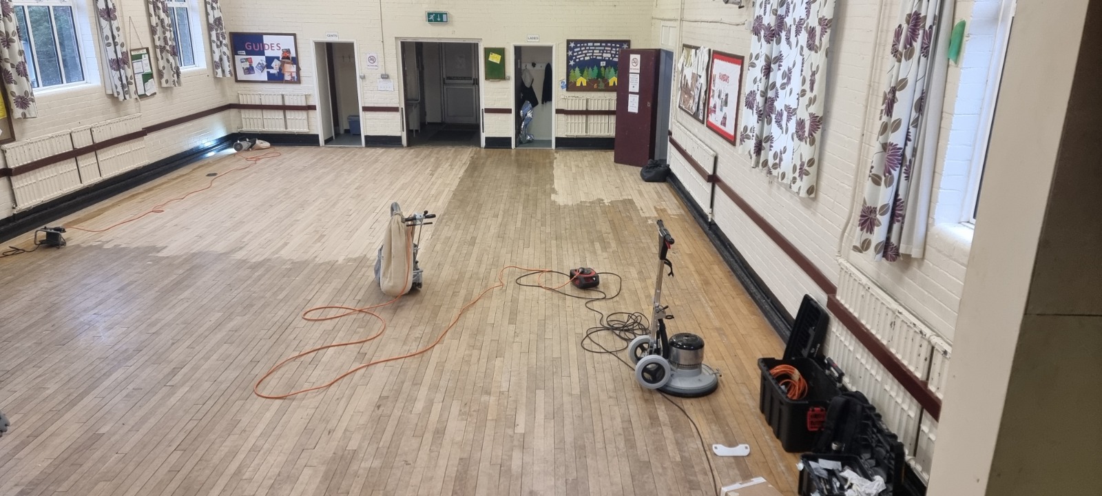 Hardwood Floor Sanding in Surbiton — community hall hardwood strip floor mid-sanding with belt sander and buffer, showing lightened lanes and preparation for refinishing.