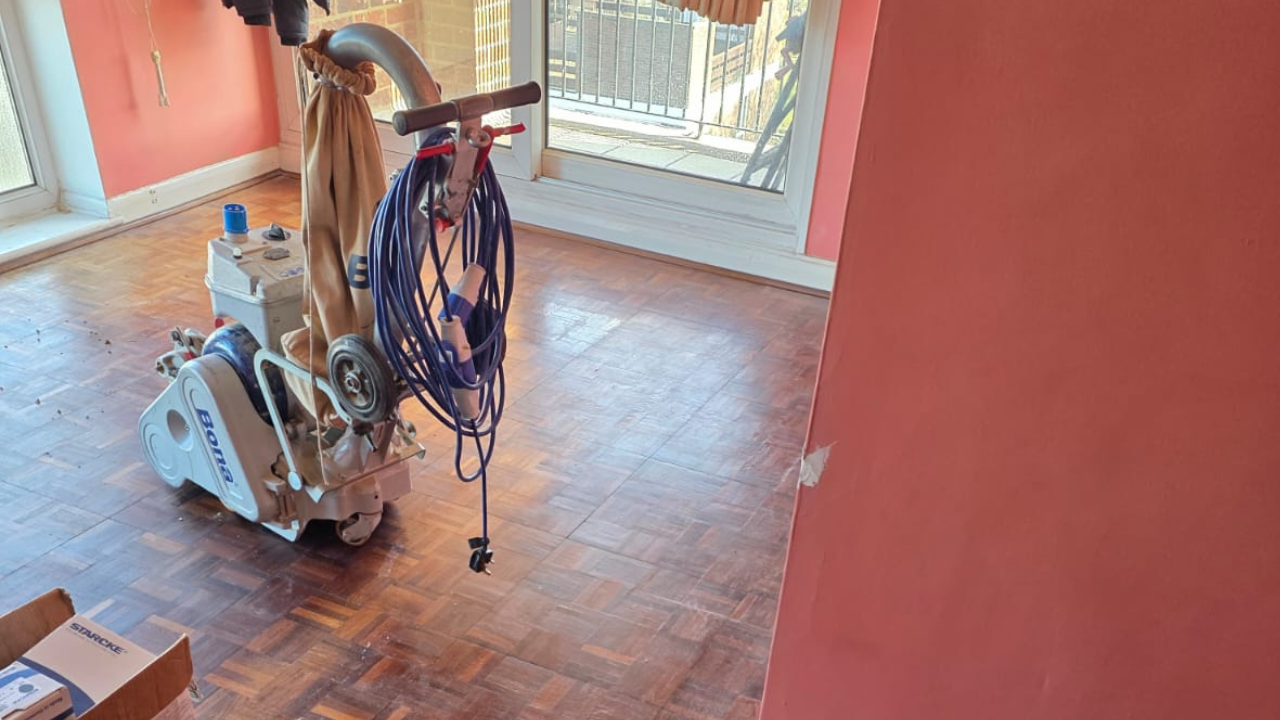 Wood Floor Sanding in – Oak Mosaic Parquet Living Room Wood Floor Sanding in on an oak mosaic parquet living room floor, with a professional dust-free sanding machine preparing the surface for restoration.