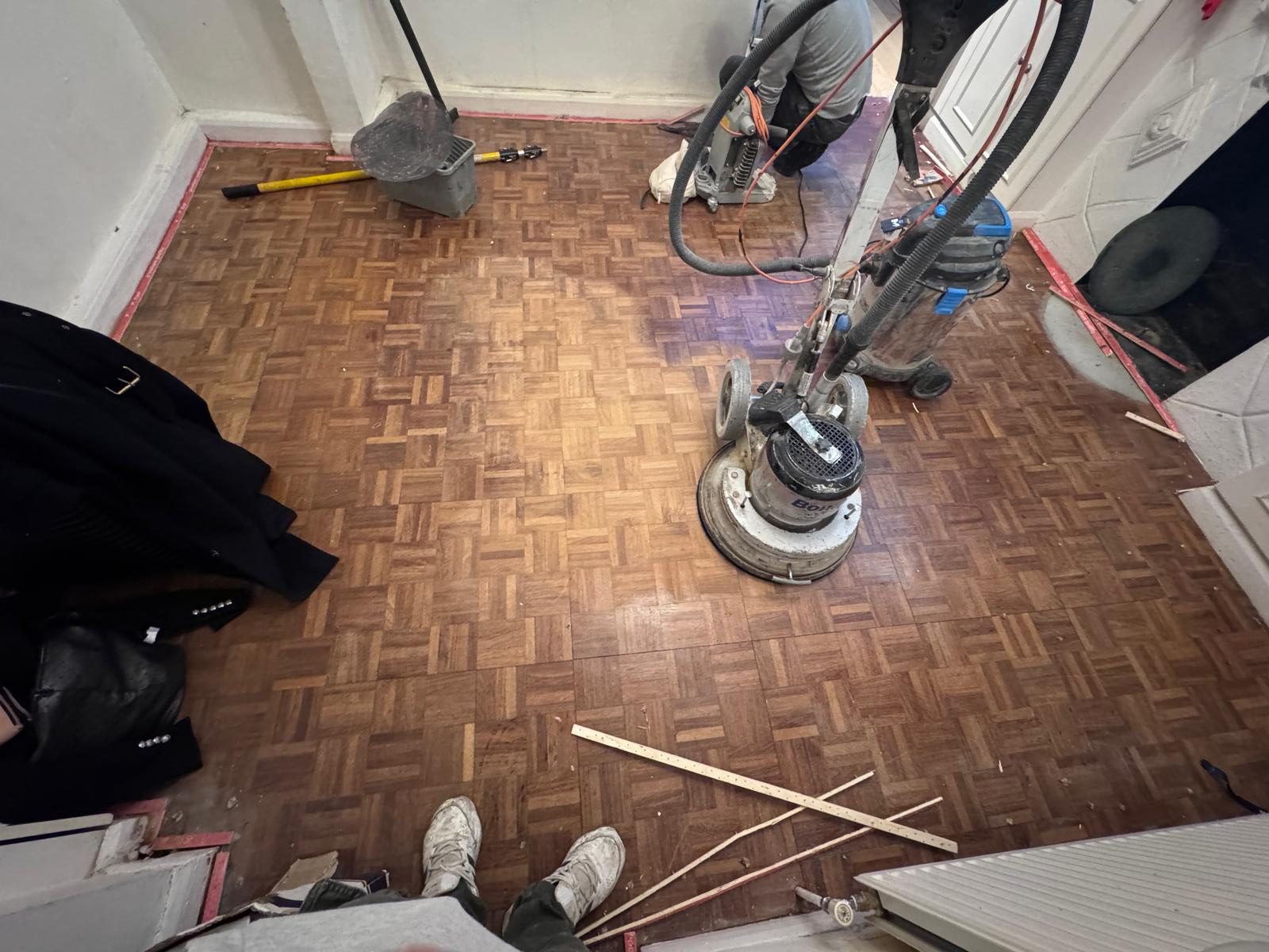 Wood Floor Sanding in Surbiton on an oak mosaic parquet hallway floor, with a rotary sanding machine and vacuum system preparing the timber surface for restoration.