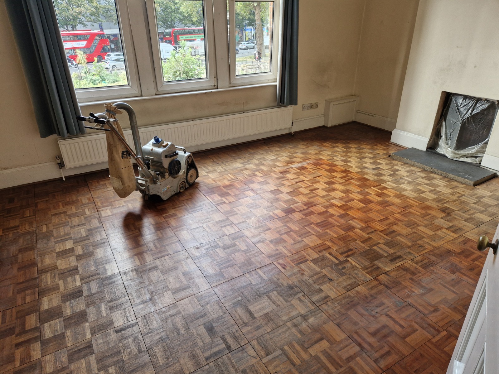 Wood Floor Sanding in Surbiton on an oak mosaic parquet living room floor, with a professional belt sander partway through restoring the worn timber surface.